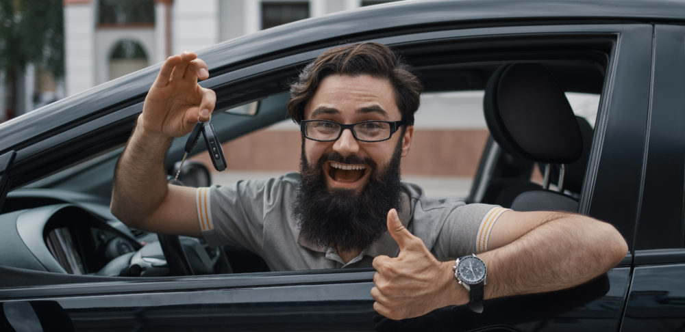 Car dealership, the happiest client. Charismatic man holding car keys showing thumbs up and smiling, with teeth, while sitting in the car looking to camera through the window..