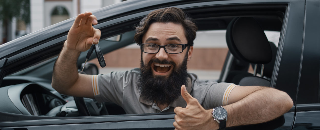 Car dealership, the happiest client. Charismatic man holding car keys showing thumbs up and smiling, with teeth, while sitting in the car looking to camera through the window..